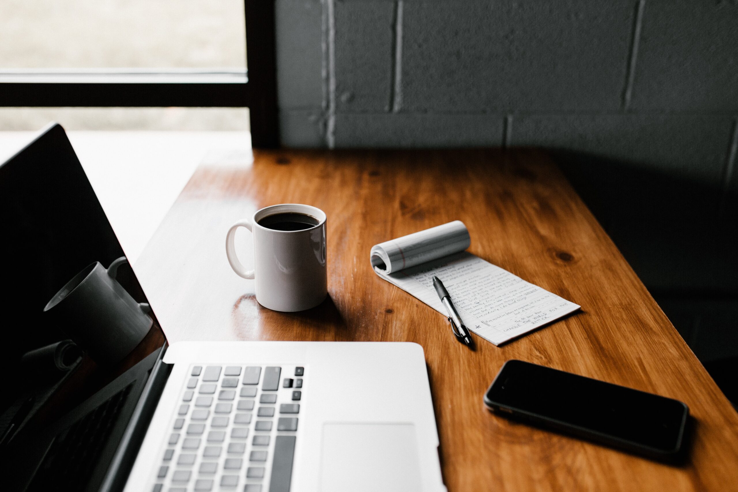 Laptop sitting next to a cup of coffee, notepad, and cell phone.