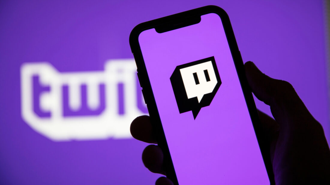 Phone with Twitch Logo being held up in front of out of focus purple Twitch logo.